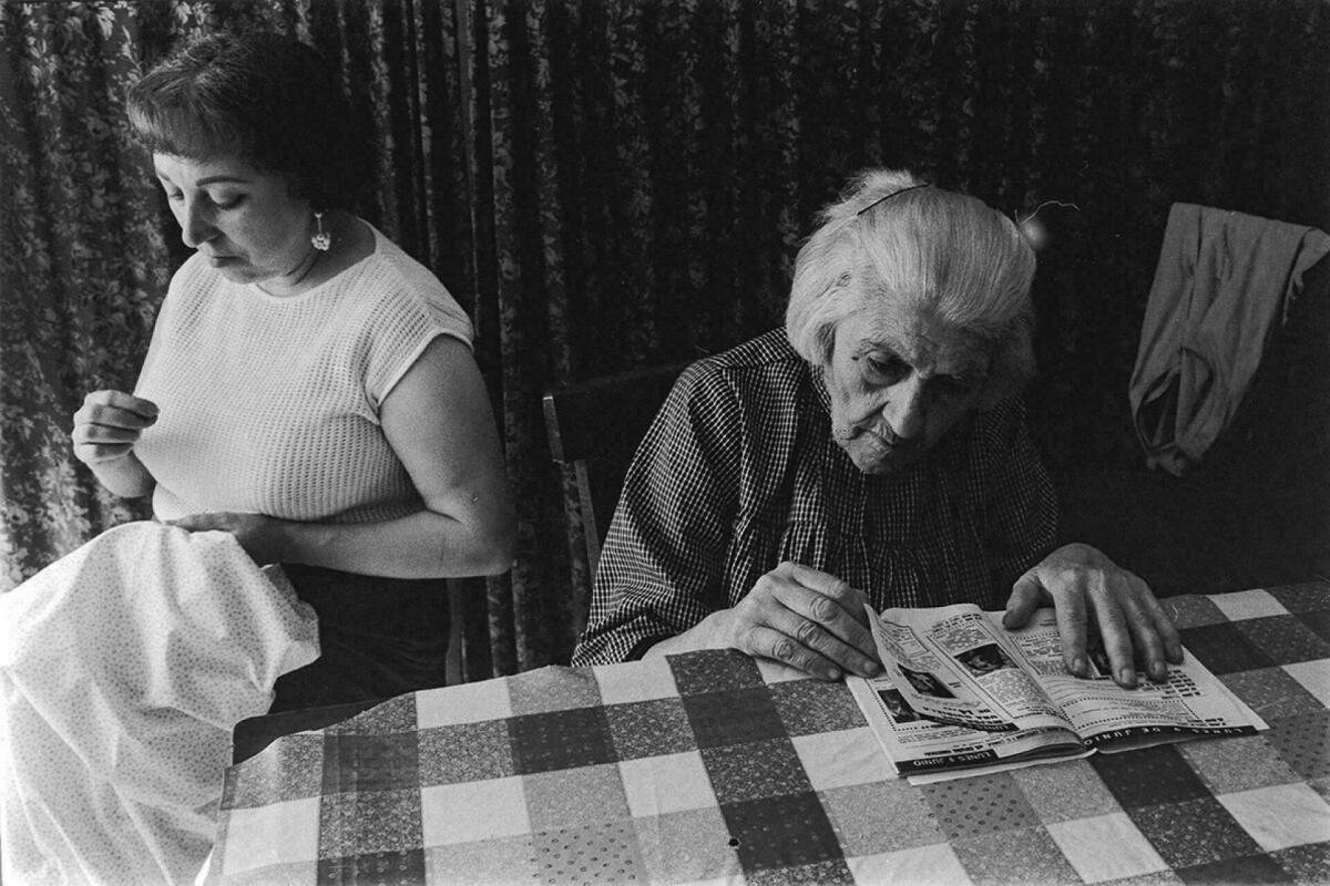Foto en blanc i negre. Es veu una escena amb una senyora cosint i una iaia llegint revista 
