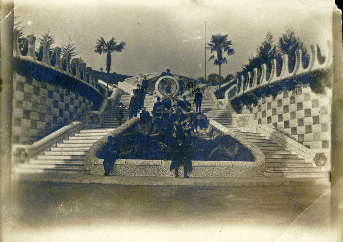 Foto en blanc i negre. Se ve la escalinata central del Parc Güell acabada de construir. Un grup de senyors estan posant  per a la foto  