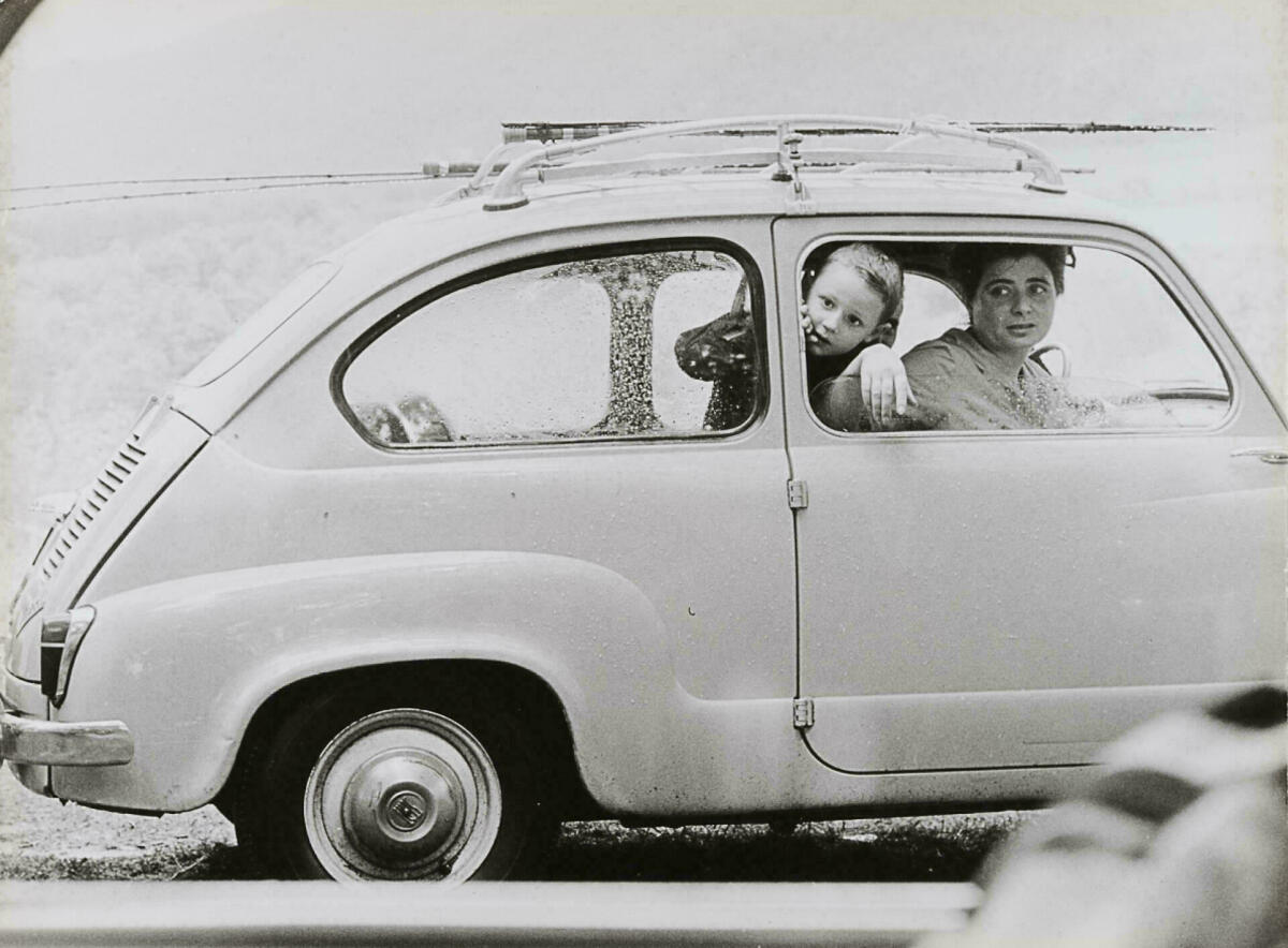 Foto en blanc i negre. Es veu un cotxe model Sis-cents amb mare i filla a dins mirant per la finestra