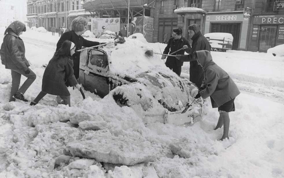 Foto en blanc i negre. Es veu un grup de 6 persones molt abrigades  treien neu de damunt de cotxe que està sepultat per la neu caiguda