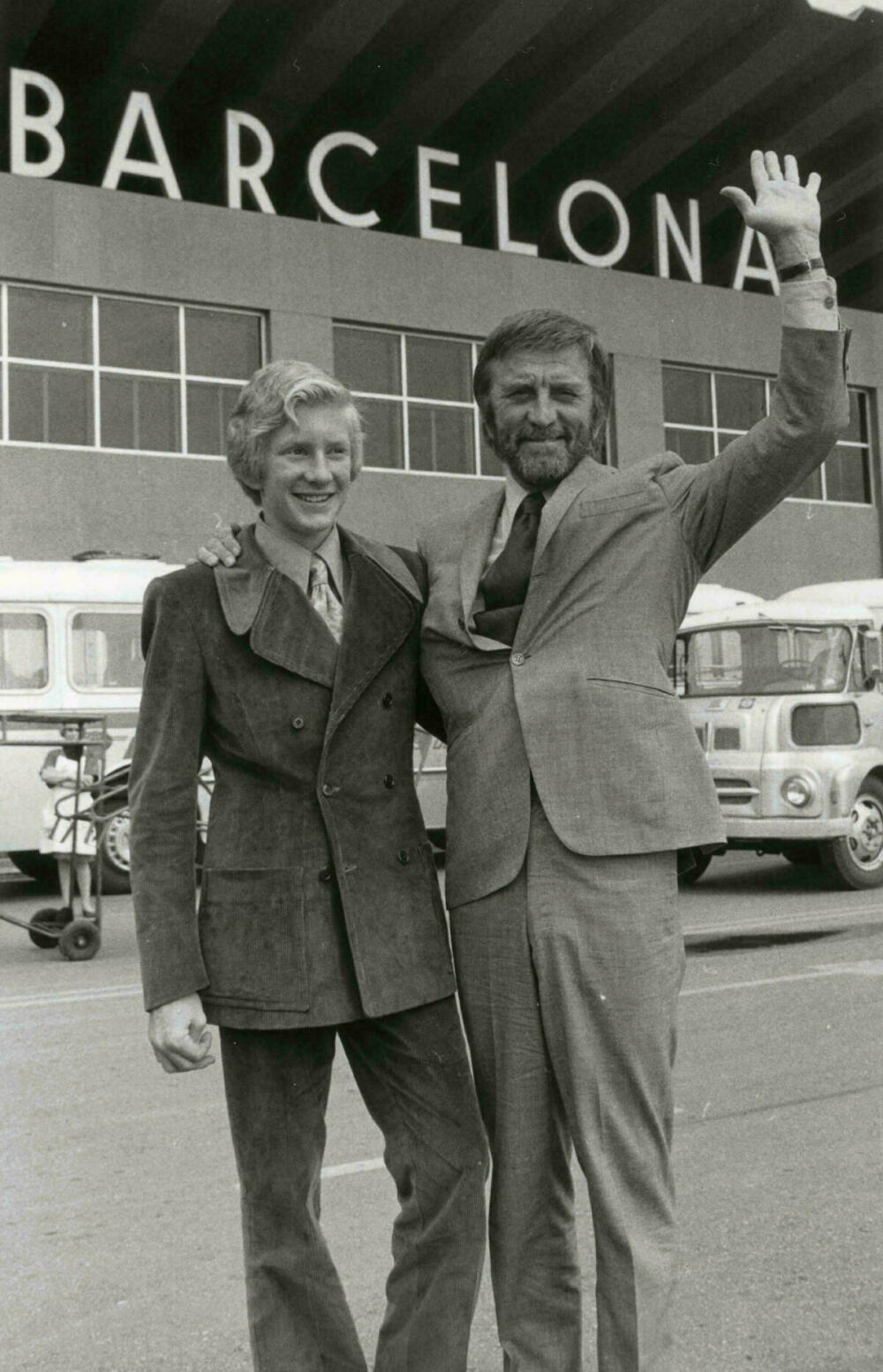 Foto en blanc i negre. Retrat de l'actor Kirk Douglas i el seu fill Peter davant l'aeroport del Prat. L'actor està saludant i somrient