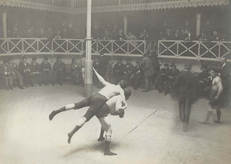 Foto en blanc i negre, públic en cercle i dos homes fent lluita grecorromana al centre
