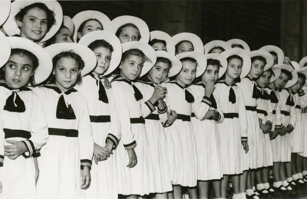 Foto en blanc i negre de nenes col·locades en fila, vestides de blanc a la processó de la Mercè