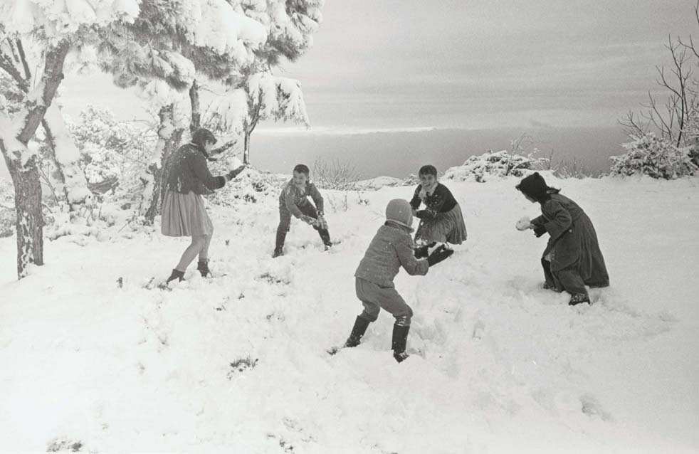 Foto en blanc i negre. Es veu un paisatge nevat amb cinc nens que juguen amb boles de neu 