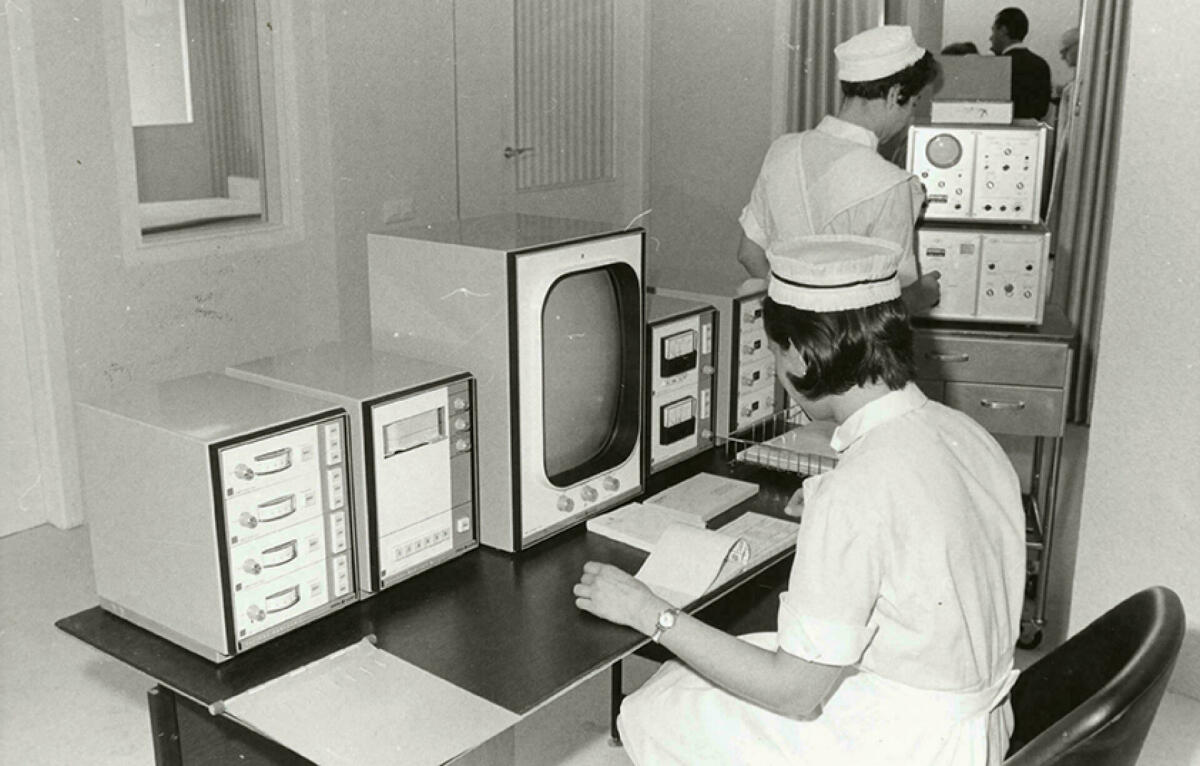 Foto en blanc i negre. Es veu un laboratori de cardiologia amb una infermera asseguda davant pantalles 