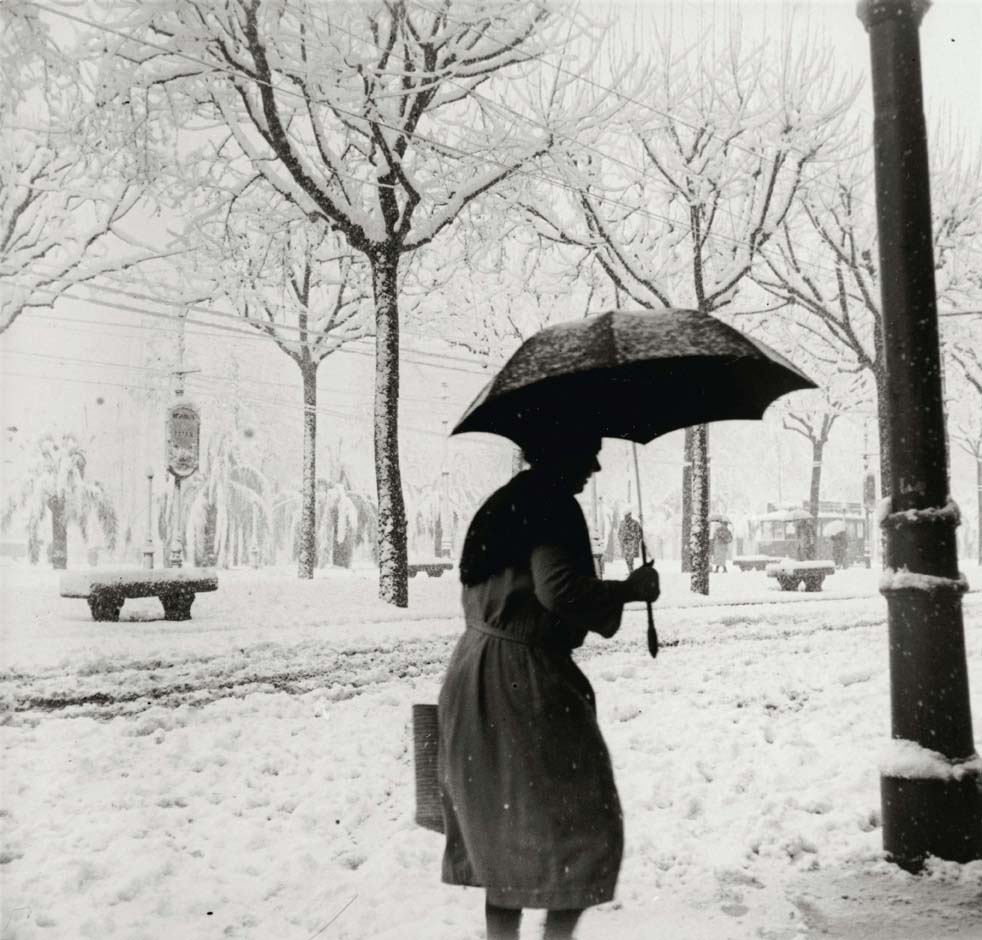 foto en blanc i negre. Es veu una vianant amb paraigües i cistell camina per un carrer ple de neu