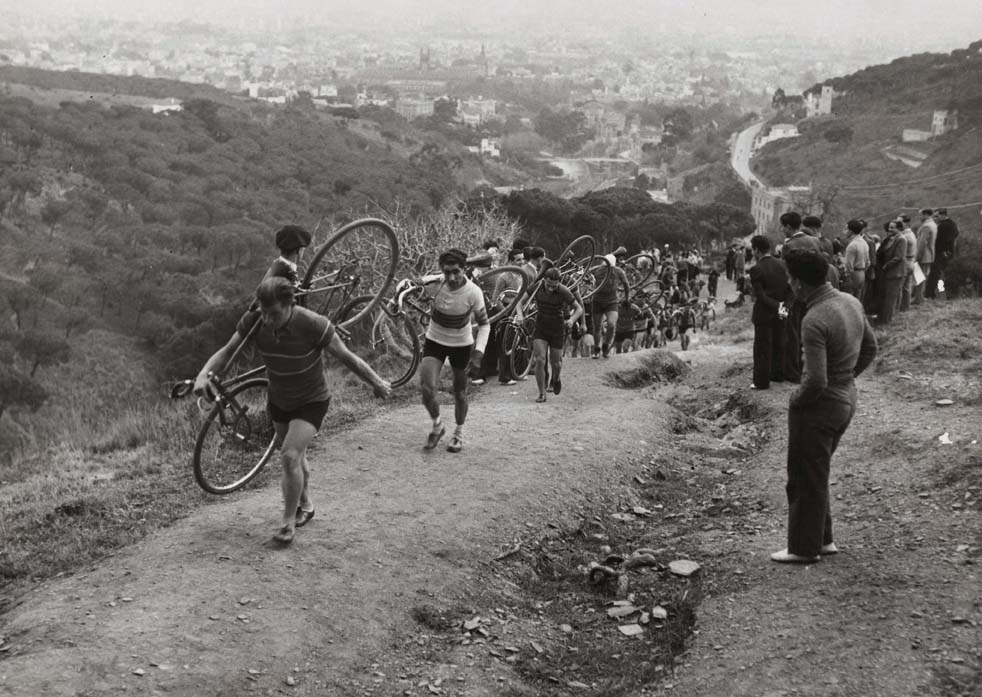 Foto en blanc i negre dels ciclistes que porten la bicicleta a l'esquena per pujar un turó