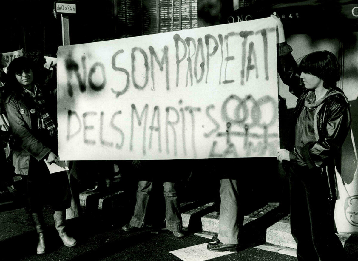 Foto en blanc i negre. Es veu 2 noies en una manifestació feminista que porten pancarta on diu : no som propietat dels marits