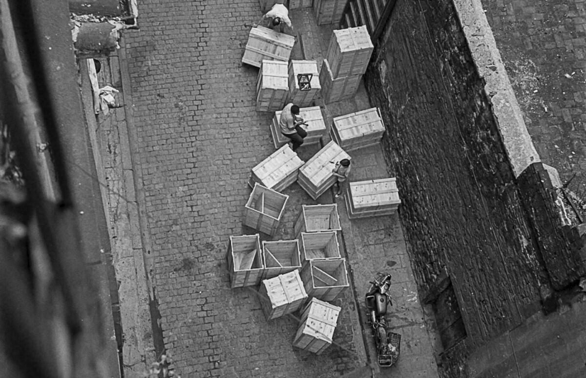 Foto blanc i negre. Es veu una vista del carrer Carabassa des del terrat  amb capses de fusta grans . Un home assegut damunt capsa i un nen jugant 
