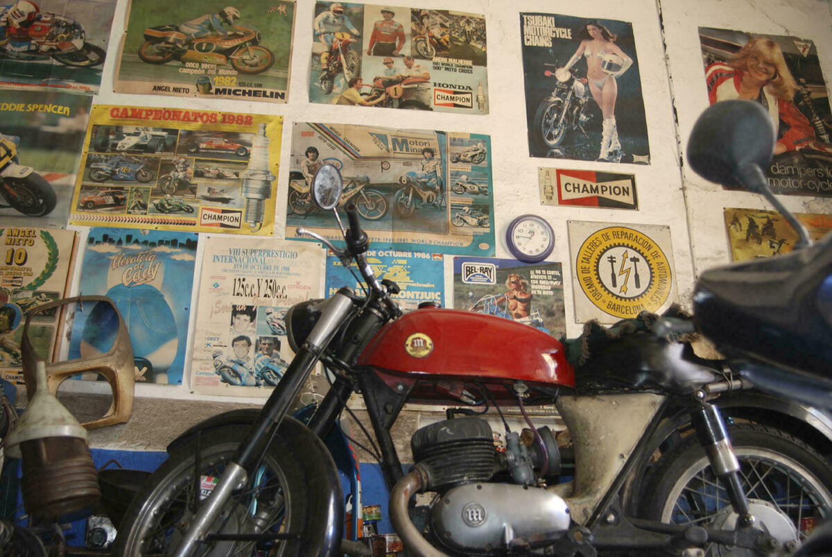 Foto a color. Es veu moto en primer terme i al darrera a la paret del taller calendaris i fotos enganxades