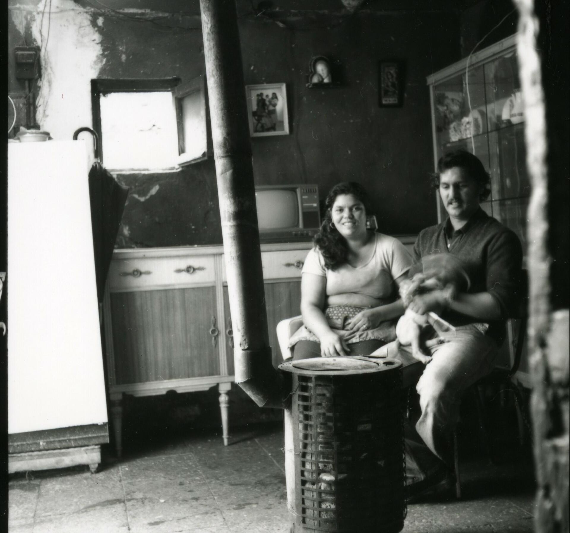 Foto en blanc i negre. Es veu linterior dun pis amb una parella somrient asseguts posant davant una estufa de llenya. al fons una cuina. 