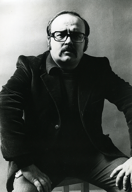 Foto en blanc i negre. Es un retrat de l escriptor Manuel Vázquez Montalbán  assegut en una cadira vestit amb americana i les mans damunt les cames