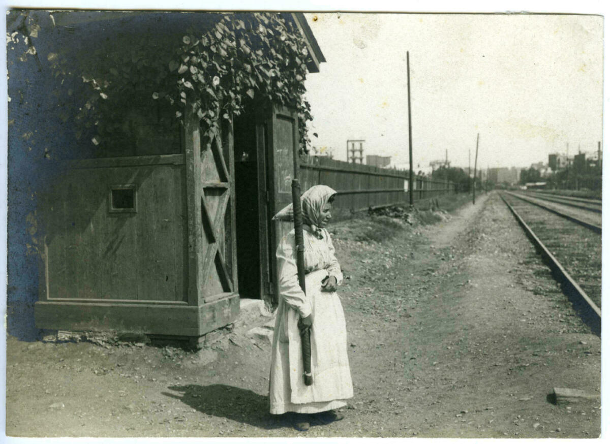 Foto en blanc i negre. Es veu dona que treballa de guardabarreres de tren davant una via