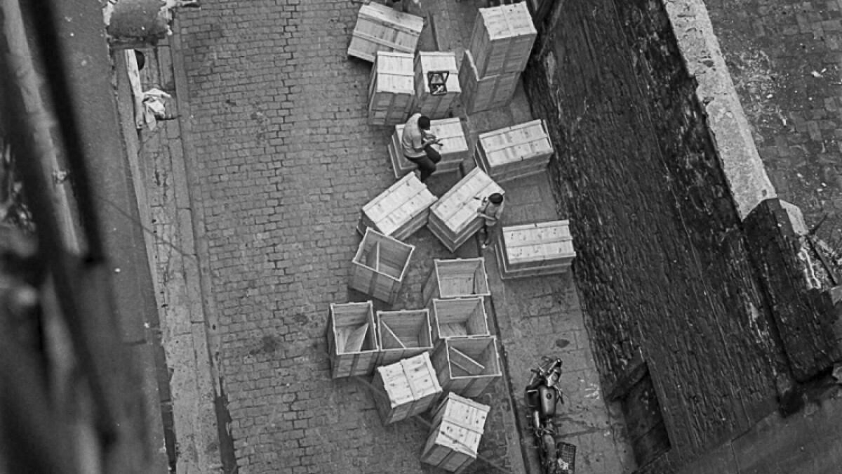 Foto blanc i negre. Es veu una vista del carrer Carabassa des del terrat  amb capses de fusta grans . Un home assegut damunt capsa i un nen jugant 