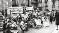 Todo un grupo bastante numeroso de niños, sentados en medio de la plaza de Sant Jaume, sostienen pancartas con lemas a favor de la escuela pública. Una niña en primer plano está tocando una guitarra y otros niños llevan las pancartas manuscritas colgadas del cuello.