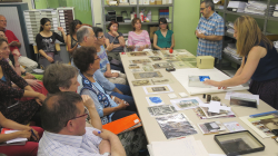 Numerous seated people listening to two people standing behind a table covered in old photos