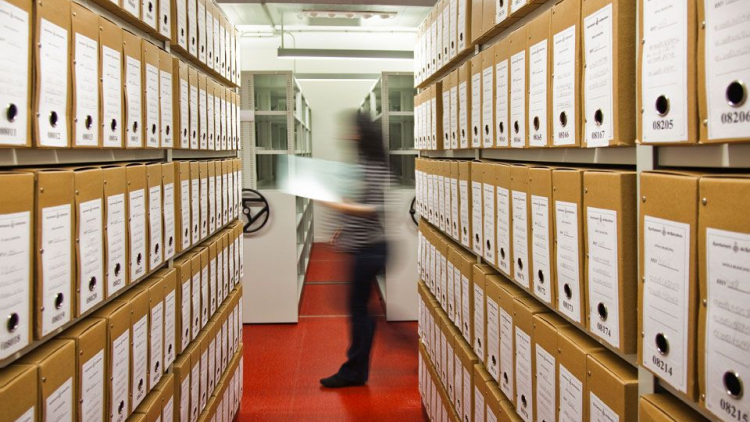 Vista d'un passadís que s'obre entre dues prestatgeries plenes de caixes d'arxiu. Al fons, es contempla la imatge d'un arxiver en moviment. 
