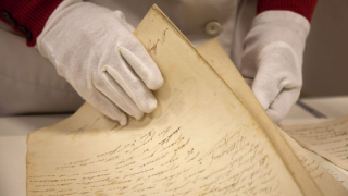 Unas manos con guantes sujetan documentos antiguos manuscritos en un archivo.
