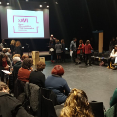 Els i les assistents a la trobada repartits en una sala amb una pantalla de fons amb el logotip de la XAVI.