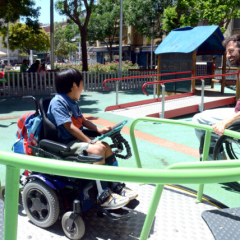 Un niño en silla de ruedas juega en una plataforma en un parque inclusivo. A su derecha, un adulto en silla de ruedas juega con él.
