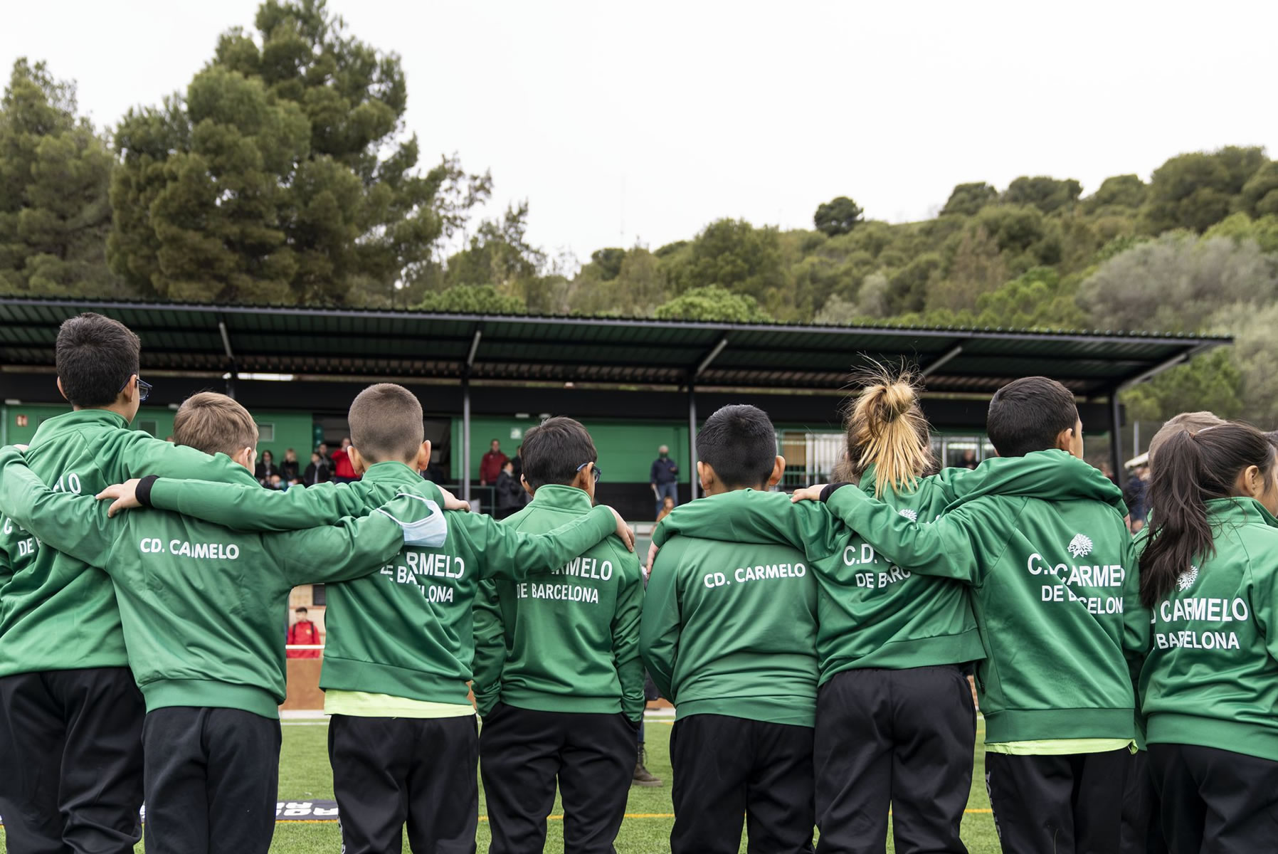 A team of child athletes with their backs turned and arms around each other looking at the football pitch