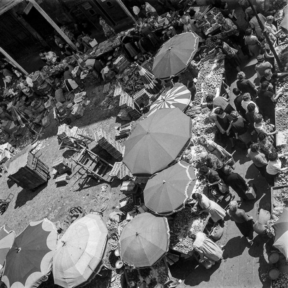 Foto en blanc i negre. Es veuen parades de fruita al carrer amb para-sols i gent