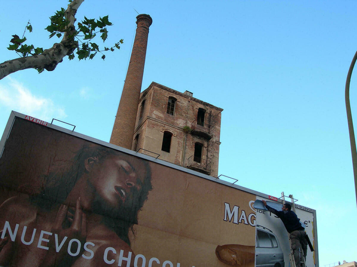 Foto a color. Es veu en primer terme una tanca publicitària amb un anunci de xocolata i al darrera la silueta d'una fàbrica amb la xemeneia