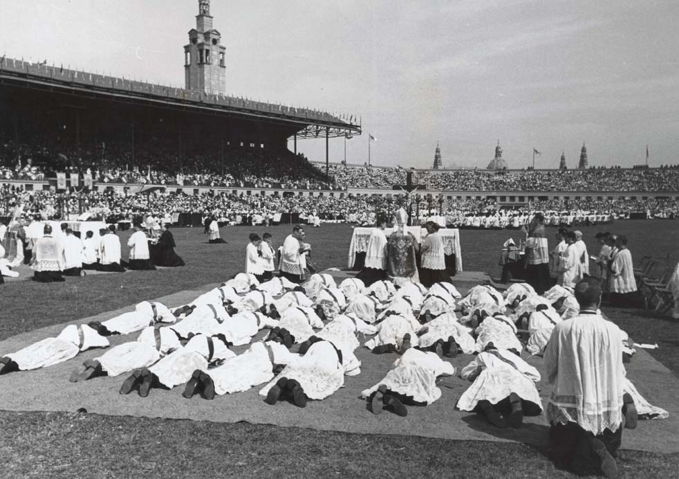 Foto en blanc i negre de la ceremonia d'ordenació de nous sacerdots estirats a terra a l'estadi de Montjuïc