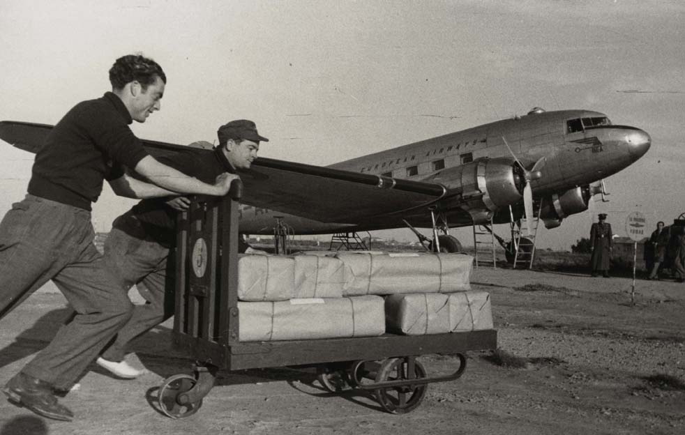 Foto en blanc i negre de dos treballadors empenyent un carro carregat de seda cap a un avió amb destinació a Dublín
