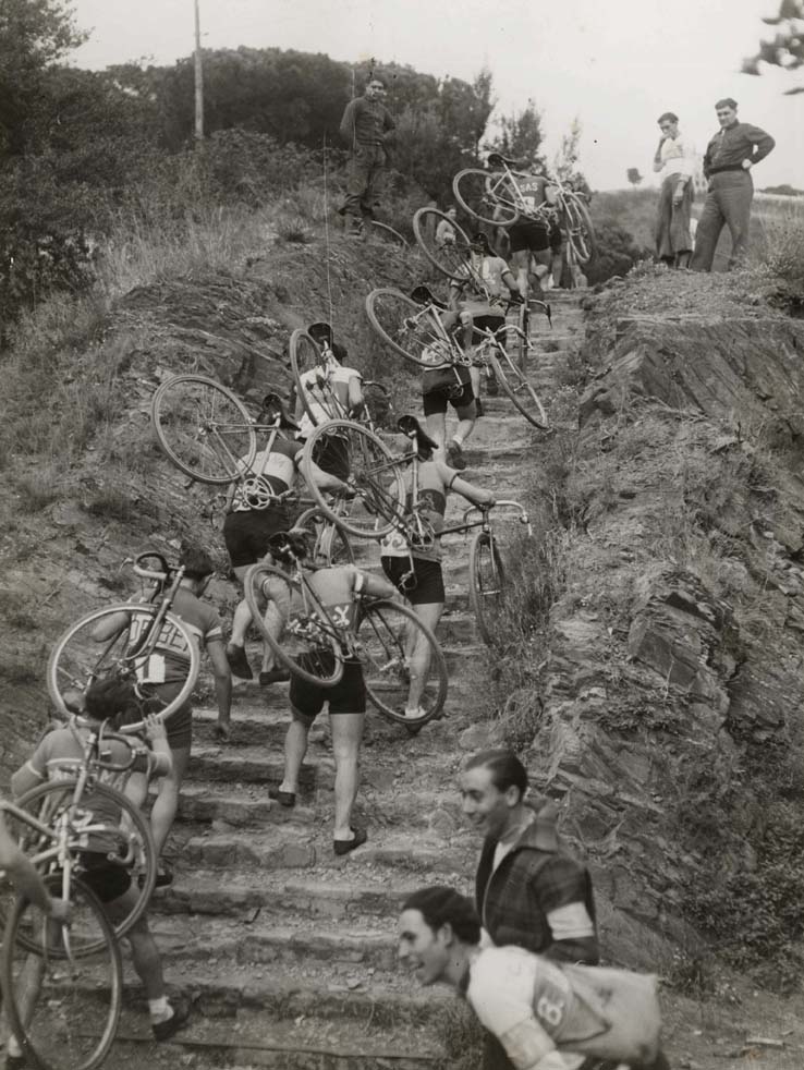 Foto en blanc i negre dels ciclistes que porten bicicletes a l'esquena per pujar un turó durant el Campionat de Catalunya per a vianants amb bicicleta. Drecera de Vallvidrera