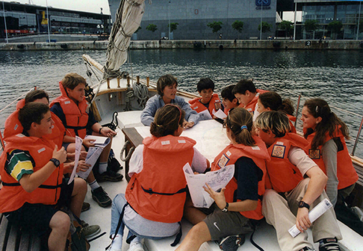 Foto color. Es veu una activitat educativa amb nens que porten armilla salvavides taronja navegant en un veler pel port.