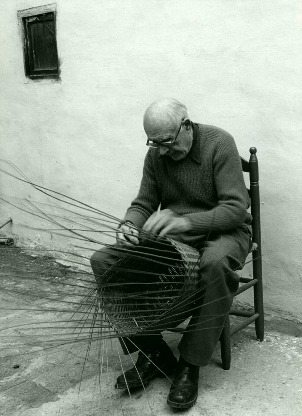 Foto en blanc i negre. Es veu un artesà assegut fabricant amb les seves mans un cistell