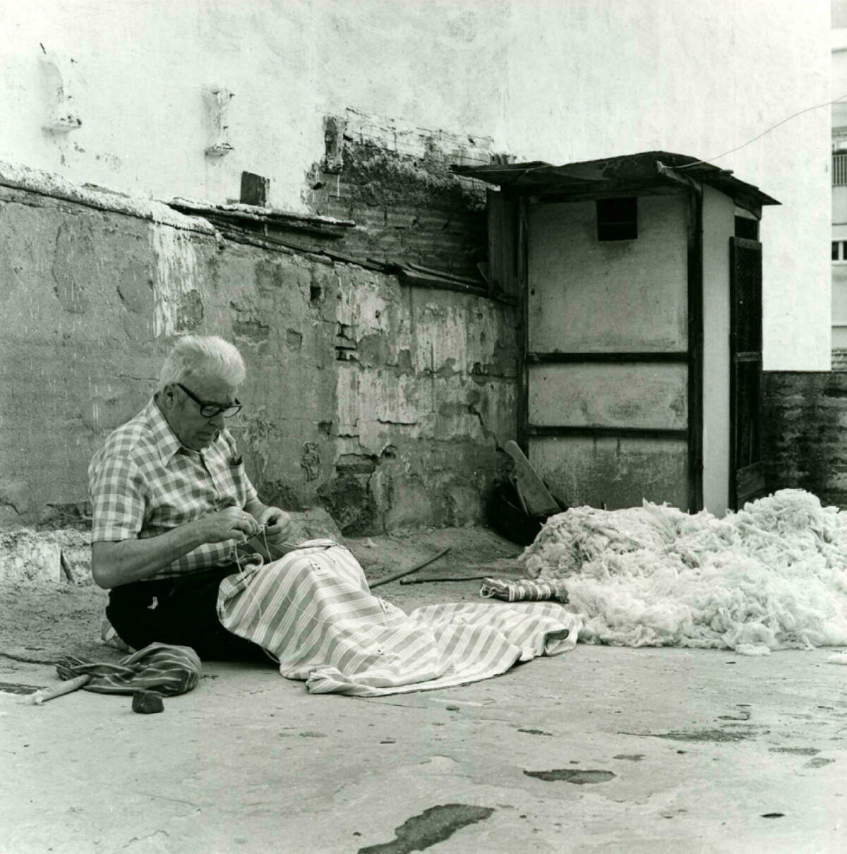 Foto en blanc i negre. Es veu un artesà assegut cosint un matalàs de llana en un terrat