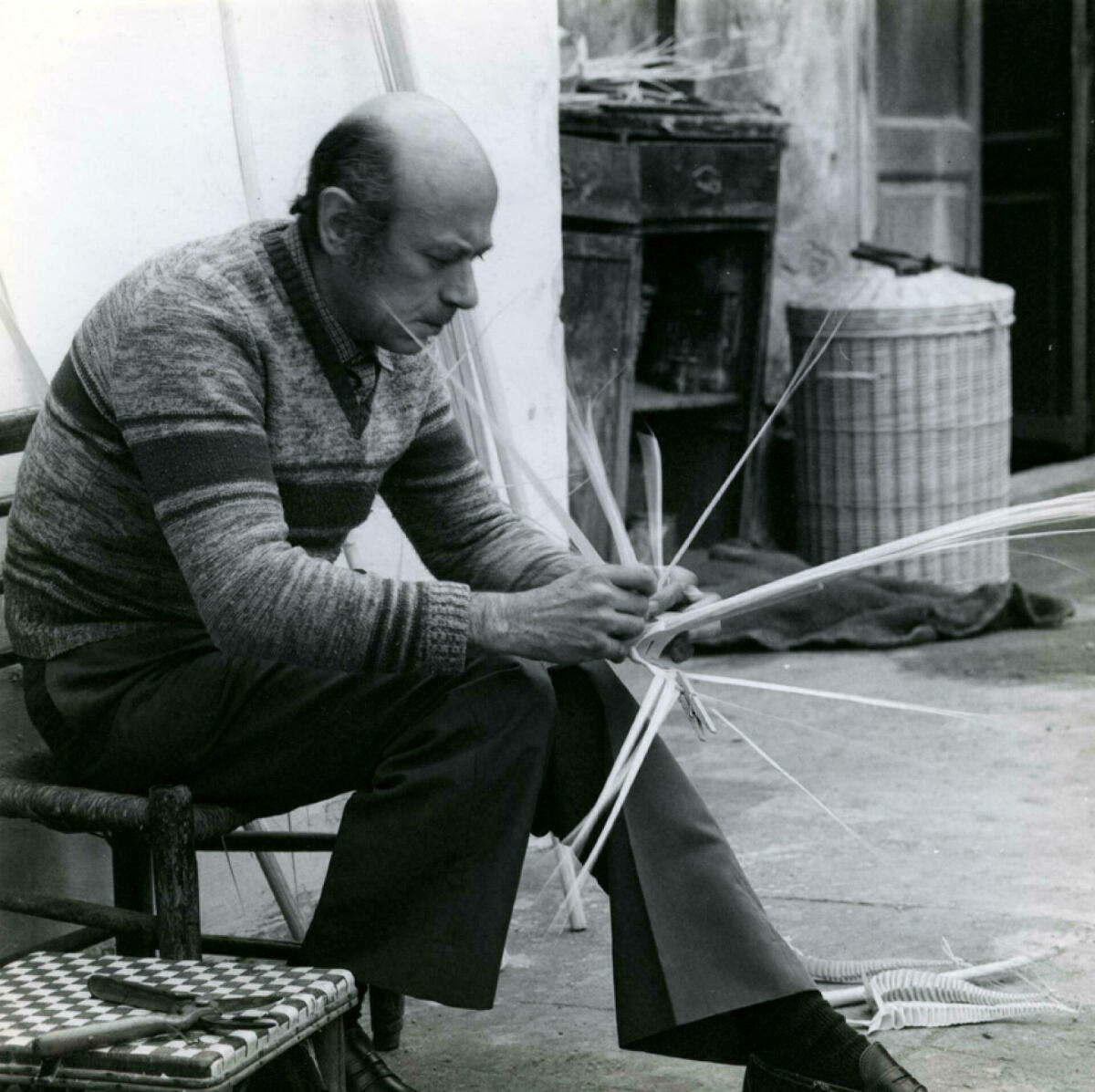 Foto en blanc i negre. Es veu un artesà palmer assegut en el seu taller fent un palmó amb les seves mans 