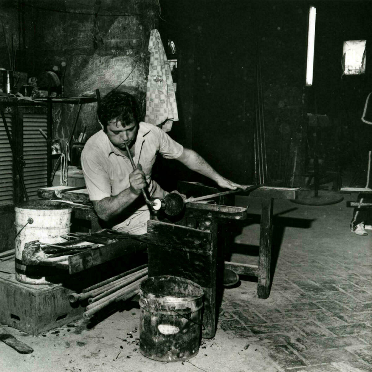 Foto en blanc i negre. Es veu un artesà vidrier assegut en el seu taller bufant vidre per fer una ampolla