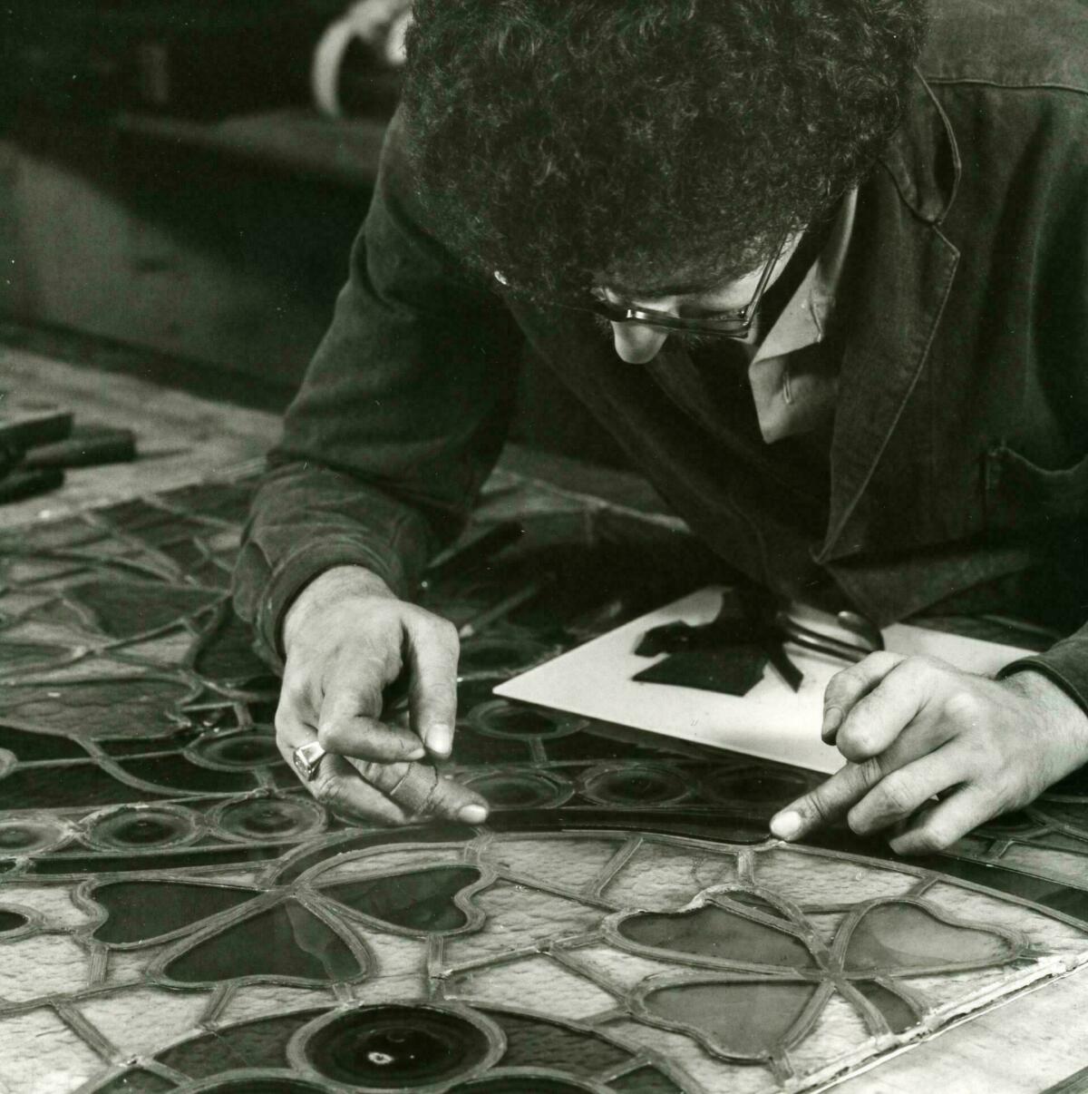 Foto en blanc i negre. Es veu un artesà vitraller assegut posant peces de vidre en un vitrall situat damunt una taula