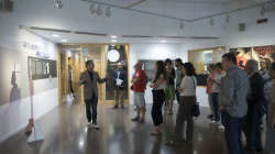 Un grup de persones escolten atentament les explicacions del comissari de l’exposició “Barcelona Coopera”. La sala està decorada amb plafons i obres que formen part de l’exposició. 