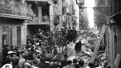 A crowd of people in the middle of a street in ruins following an air raid. 