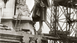 In the picture, you can see how some workers are getting ready to raise the Christopher Columbus statue with pulleys to the top of the monument dedicated to him. To the right, there is an iron structure with ladders acting as a support for the general construction of the monument, while in the foreground, there is a worker with a portable ladder. 