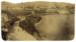 Picture of Barcelona from the mid-19th century, where you can see the walls which follow the contours of the first city, the one that opened out to the sea. The figures of three people walking along the coastline appear in the foreground.
