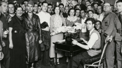 A large crowd of people surround three couples getting married at the same time, in front of a secretary who is sitting down and typing. Everyone is looking at the camera for the photo. 
