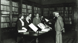 A black and white photograph showing a room with shelves full of books and a table with books on display; several people at one side of the table are looking at two others consulting a book.