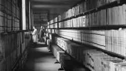 A black-and-white photograph where you can see shelves full of old books on the right and two people in the background. One of them is very close to a window through which natural light enters for consulting the documents.