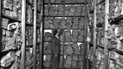 Fotografía en blanco y negro que muestra estantes llenos de volúmenes de documentos antiguos con una figura con bata gris en medio que señala uno de los volúmenes.