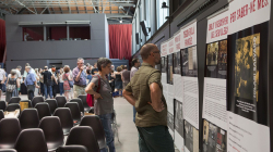 A group of people carefully reading the explanations on some exhibition panels. In the background, you can see more people waiting for the conference to start and, in the foreground, empty chairs ready for participants to take their seats. 