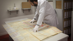 A person with a white smock and gloves flattens out a map on a table in a room with shelves full of boxes