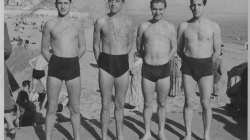 Four men in swimming trunks stand behind bathers, beach and seashore, with the buildings of Barceloneta in the background. 1946. Claret. La Barceloneta People’s Archive Collection. AMDCV