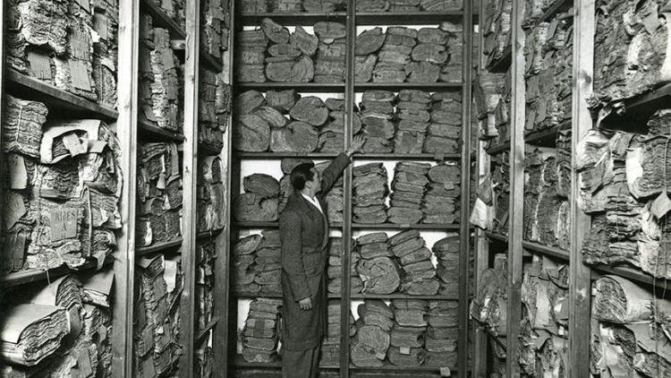 Fotografía en blanco y negro que muestra estantes llenos de volúmenes de documentos antiguos con una figura con bata gris en medio que señala uno de los volúmenes