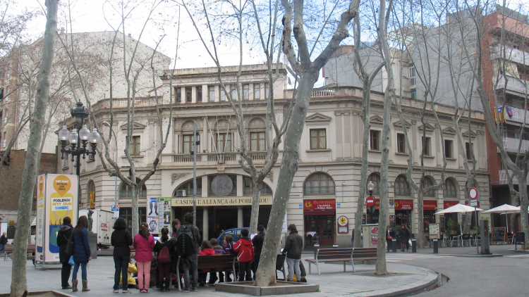 Un grupo de alumnos de Primaria se encuentran rodeando un educador mientras escuchan sus explicaciones. Todo el grupo se encuentra en medio de la plaza frente al Casino de la Alianza del Poblenou.