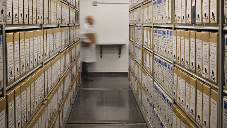 At the end of a corridor, boxes of records can be observed on either side and the figure of an archivist.
